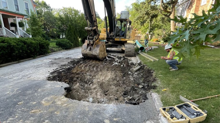 Worker in large backhoe removing asphalt from a road with workers and tools around it