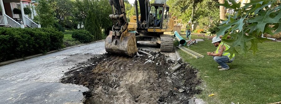 Person in backhoe digging large hole into asphalt pavement with people and tools around it