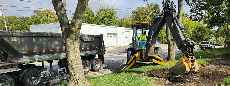 Person in backhoe digging up dirt where a sidewalk will be installed. A tractor trailer is parked to the left of the backhoe.