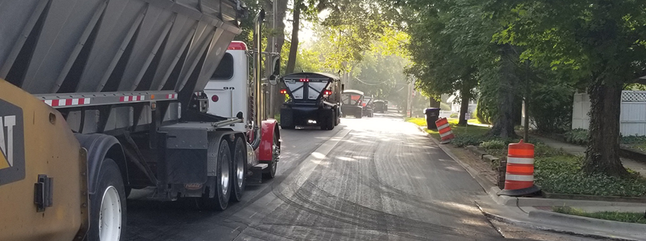 Several asphalt trucks driving down a road with a milled service and traffic barrels on the side