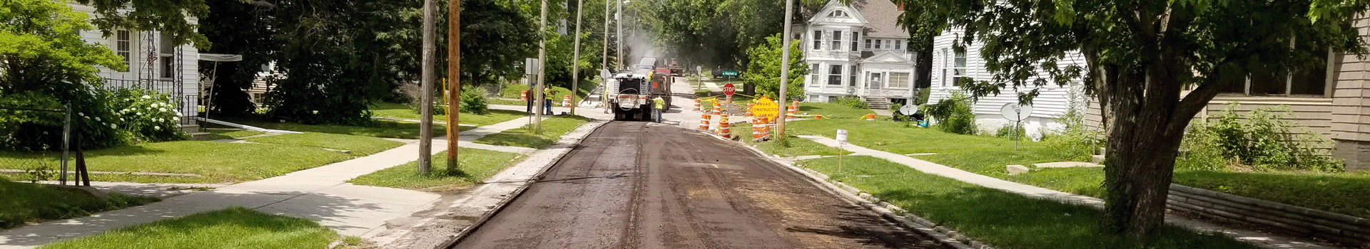 Long distance photo of road with the top surface mostly removed. Milling equipment, workers, and cones are at an intersection with milling operations in progress