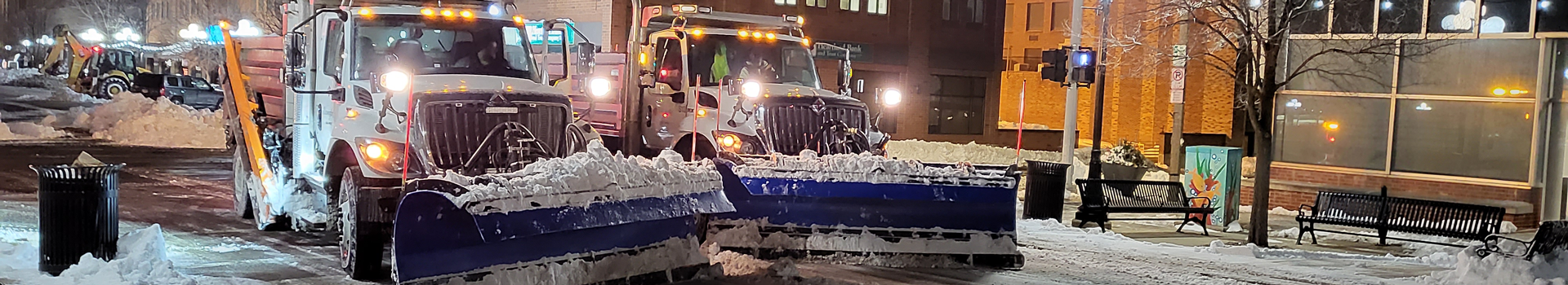 Two snow plows next to each other on a street with snow cleared in front of them and snow on the sides